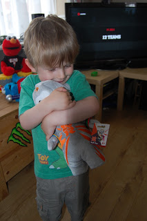 A young child with blond hair hugs a grey and orange Wenlock soft toy, standing in a living room. An Elmo plush is on the sofa, and a TV in the background displays the words "FILMS" and "12 YEARS.