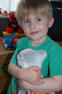 A young child with blonde hair and blue eyes, wearing a green shirt, smiles whilst hugging a grey Wenlock soft toy in a playroom with toys visible in the background.