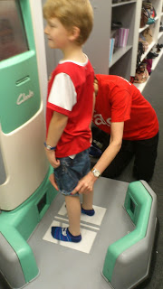 A young boy stands on a foot measuring device as a shop assistant adjusts his position. Both in red shirts, they're preparing for Back To School season at a shoe shop, with shelves of Clarks school shoes visible in the background.