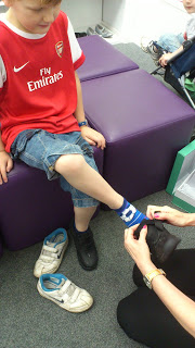 A child in an Arsenal football shirt and blue shorts sits on a purple bench as an adult helps him try on a black Clarks shoe—perfect for that Back To School moment; his other shoe and socks rest on the floor.