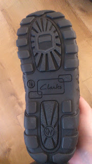 The image shows the underside of a black Clarks school shoe, highlighting its textured rubber sole with deep treads and the brand name "Clarks" embossed in the centre. Perfect for Back To School, a wooden floor is visible in the background.