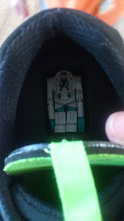 A small toy robot is placed inside the opening of a black Clarks shoe with a green pull tab. A person's finger holds the shoe open, highlighting a fun Back To School moment.