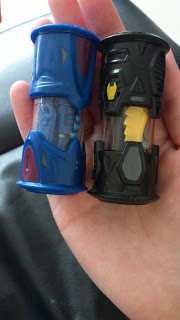 A hand holding two small toy capsules, resembling Monsuno Cores: one blue with a purple accent, and one black with yellow and grey accents. Each capsule has a tiny dinosaur figure inside, ready for your next Monsuno Strike.