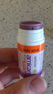 A hand holds an open tube of Nelsons Arnicare kids stick with a purple balm—one of the mummy first aid must-haves—against a light-coloured, speckled background.