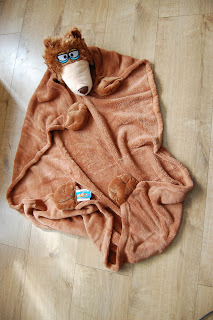 A soft brown CuddleUppet blanket with a bear's head, wearing blue glasses and bear paw details, is spread out on a wooden floor—perfect for cosy snuggles.