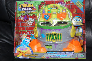 A boxed toy set called "The Trash Pack," featuring a colourful rubbish-themed playset with slime designs, orange ramps, and small character figures like Scum Drum, displayed against a brick wall backdrop inside the packaging.