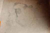 A beige carpet with several large, dark, irregular stains scattered across its surface—perfect for demonstrating the power of a Rug Doctor carpet cleaner.
