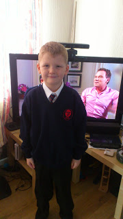 A young boy in a school uniform stands smiling in front of a TV, which displays a man in a pink shirt sitting on a sofa. The scene captures the feeling of back to normality, with light walls and some furniture visible in the room.