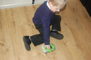 A young boy in a dark jumper and black trousers sits on a wooden floor, playing with Lex the T-Rex from VTech’s Switch & Go Dinos collection.