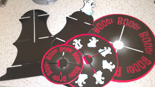 Three black plastic pieces from Poundland with slotted sections for assembly. Two round pieces say "BOOO!" in red with ghost illustrations, and one bat-shaped piece features a small ghost—perfect for Halloween décor. All are on a light worktop.