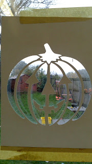 A sheet of paper with a Poundland Hallowe’en pumpkin and anchor pattern cut out, taped to a window. Through the cut-outs, a garden with grass, a shed, and outdoor toys is visible.