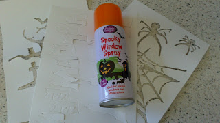 A can of Spooky Window Spray from Poundland, with an orange lid, sits atop two Hallowe’en-themed stencils featuring bats, spiders, and cobwebs on a speckled worktop.