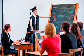 A child wearing a graduation cap and gown stands at a blackboard, pointing at a diagram, while adults seated at desks use tablets, highlighting how technology plays a role in modern parenting and children's education.