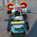 A colourful LEGO vehicle model with orange wheels, a transparent blue windscreen, and various grey, yellow, and black bricks—just like something from The Lego Movie—displayed on a tiled floor.