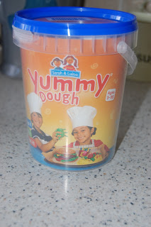A plastic container of "Yummy Dough" with a blue lid sits on the kitchen worktop, featuring two kids in chef hats. Perfect for fun activities and kids baking, the label reads "Sarah & Luba’s Yummy Dough, Ages 3-99.