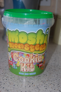 A plastic tub labelled "Moshi Monster Cookie Kit" with a green lid sits on a worktop. The colourful packaging features cartoon monster characters, making this Cookie Kit perfect for fans of Moshi Monster treats.