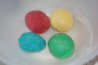 Four colourful dough balls—red, yellow, blue, and green—sit in a white mixing bowl. Perfect for children's cooking or fun activities, these Yummy Dough balls are slightly dusted with flour.