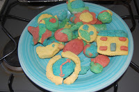 A blue plate on a cooker holds various colourful, hand-shaped biscuits made from tasty dough—shaped like a flower, house, clock, and more—perfect for children's baking and fun activities.