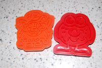 Two colourful plastic biscuit cutters shaped like Moshi Monster cartoon characters, one orange and one red, placed on a speckled worktop—perfect for any Biscuit Kit.