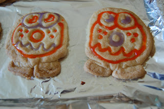 Two sugar biscuits on foil, decorated with orange and purple icing from a Biscuit Kit to resemble cartoonish Moshi Monster faces with big eyes, round noses, and jagged mouths.