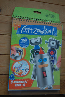 A spiral-bound Artzooka! Clingzooka activity pad titled "Clingzooka Robots" features cartoon robots and a hand placing robot stickers. Designed for ages 5+, it includes 250 sticker pieces, all displayed on a light wooden surface.