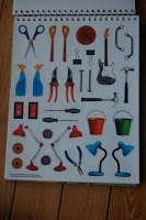 A page from a spiral-bound book showing illustrations of various objects, including scissors, spray bottles, buckets, lamps, a basketball, a guitar, tools, and even playful robots on a wooden surface.