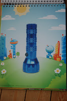 A blue torch stands upright on grass in the centre of a colourful illustrated background with cartoon buildings, white flowers, a bright sun, and playful Artzooka designs.