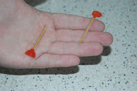 A hand holds two cocktail sticks, each skewering a small red object, possibly sweets or fruit, over a speckled worktop—perfect for a fun Artzooka-inspired snack creation.