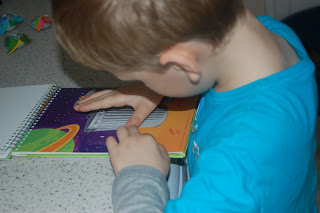 A young child wearing a blue shirt uses their fingers to read a braille book with colourful illustrations of planets and playful Artzooka designs on a table.