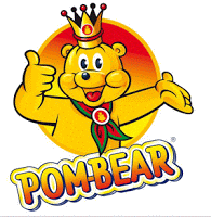 A smiling cartoon bear wearing a crown and green scarf gives a thumbs-up with one paw and gestures with the other. The word "POM-BEAR" is displayed in colourful, bold letters below, celebrating tasty Pom Bear snacks like Pigs in Blankets.