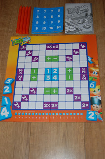 A Mathable Junior board game is laid out on a wooden floor with a colourful board, blue number tiles, orange holders, a blue score card, and an instruction booklet visible at the top.