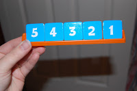 A hand holds an orange tray with five blue square tiles displaying the numbers 5, 4, 3, 2, and 1 in white—resembling pieces from the Mathable Junior Game. The background is out of focus.