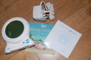 A Gro Clock sleep trainer for sleep training, a power adaptor, the children’s book "Sleepy Farm," and an instruction manual are displayed on a wooden floor—perfect for a Gro Clock review.