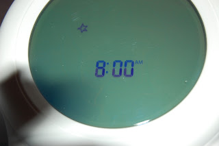 A close-up of a Gro Clock display shows the time 8:00 am in blue numbers on a green screen, with a small star icon on the left—perfect for sleep training routines.