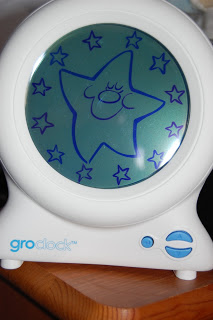 A white Groclock sleep trainer, perfect for sleep training, features a display with a large, smiling, sleeping star surrounded by smaller stars on a blue-green background. The device rests on a wooden surface.