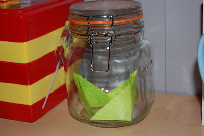 A clear glass jar with a metal clasp contains a green origami paper boat, symbolising positive thinking. The jar sits on a wooden surface next to a red and yellow striped container.