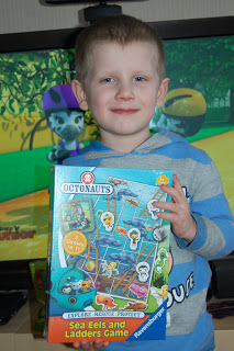 A smiling young boy in a blue striped hoodie holds up the Octonauts Sea Eels and Ladders game box. A colourful animated children’s programme is playing on the television in the background.