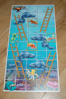 A snakes and ladders game board with an underwater theme inspired by the Octonauts Sea Eels and Ladders game, featuring sea animals like fish, octopus, turtle, and seahorses on numbered squares atop a wooden surface.