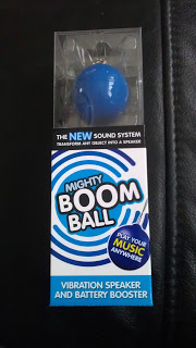 A packaged Mighty Boom Ball vibration speaker and battery booster, with a blue and white striped design and the slogan "Turn any object into a speaker," is displayed on a black surface. This portable speaker delivers powerful sound anywhere.