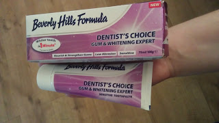 A hand holds a box and tube of Beverly Hills Formula Dentist's Choice Toothpaste & Whitening Expert Sensitive Toothpaste, featuring purple and white packaging with product claims, against a wooden floor background.