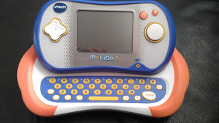 A VTech MobiGo 2 handheld educational game device with a touchscreen, round buttons, and a slide-out yellow QWERTY keyboard rests on a black surface—perfect for your next VTech MobiGo review.
