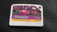 A white Vtech MobiGo game cartridge featuring characters from the film Cars, with the MobiGo and VTech logos on a purple and yellow label, lies on a black textured surface—ideal for your Vtech MobiGo 2.
