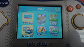 A close-up of a VTech MobiGo 2 screen shows user "james" with icons for Cartridge, Hamster Highway, Shooting Gallery, Downloads, Art Master, and Photos. The device’s buttons frame the screen in this detailed VTech MobiGo 2 review image.