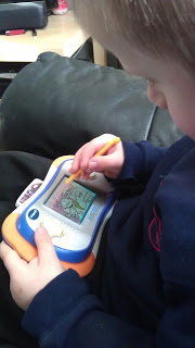 A young child is sitting on a sofa, holding a Vtech MobiGo 2 and using a stylus to colour a picture on the screen. The child's face is partially visible in profile, highlighting quiet focus—perfect for any MobiGo 2 review.
