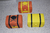 Three small, colourful treasure chests sit on a speckled surface—perfect for a Pirate Party. Two are orange with black skull-and-crossbones designs, and one is yellow with black stripes and a yellow lock symbol on the front.