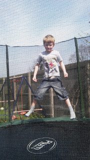 A young boy wearing a grey T-shirt and shorts is jumping on a trampoline outdoors, surrounded by a safety net, as “Here Comes The Sun” plays softly in the background, with a wooden fence and climbing frame behind him.