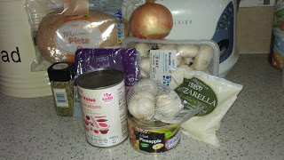 A selection of pizza ingredients on a kitchen worktop, perfect for Foodie Fun, including wholemeal pitta bread, an onion, mushrooms, mozzarella cheese, garlic, mixed herbs, a tin of tomatoes, and pineapple chunks.