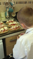 A young foodie in a white shirt stands in the kitchen, having fun as he adds toppings to slices of bread with tomato and cheese—mini pizzas arranged on a wire rack. Mugs hang on hooks above the worktop, ready for a tasty McCain’s snack break.
