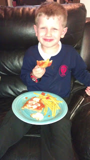 A young boy in a navy blue school uniform sits on a black leather sofa, smiling while holding a slice of pizza. A blue plate with pizza and McCain’s chips rests on his lap, adding to the foodie fun.