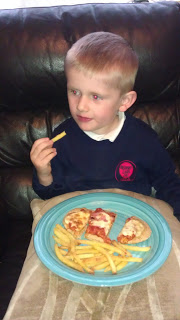 A young boy sits on a dark brown leather sofa, enjoying some Foodie Fun. Holding a chip from his blue plate of McCain's chips and pizza slices, he looks off to the side in his blue shirt with a pink logo.